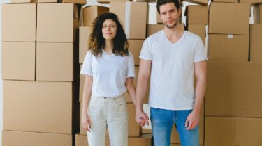 A young couple holding hands in front of a stack of cardboard boxes, possibly indicating moving or relocation.